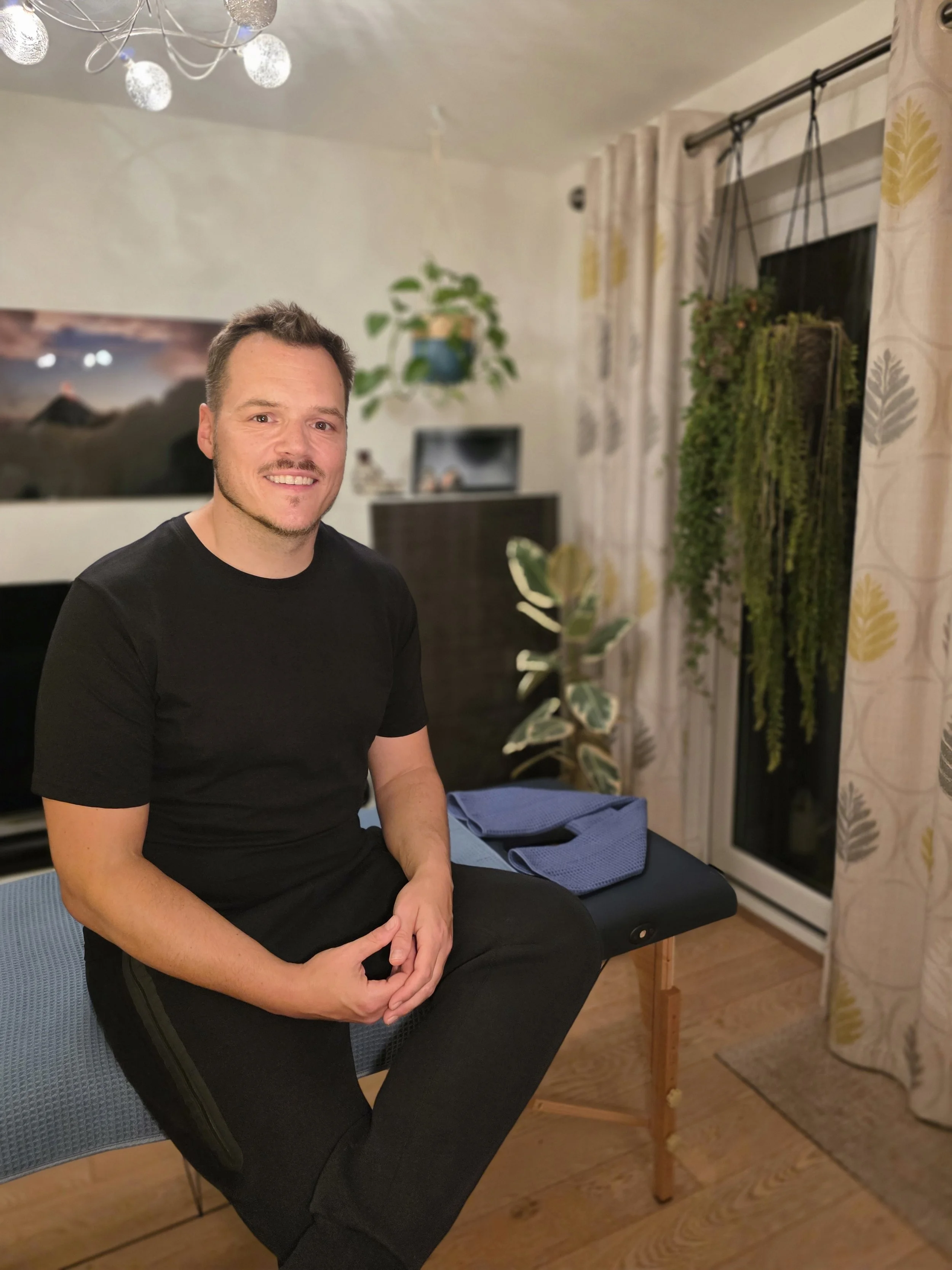 A man sitting on a massage table in a room with potted plants, curtains, and a television in the background.