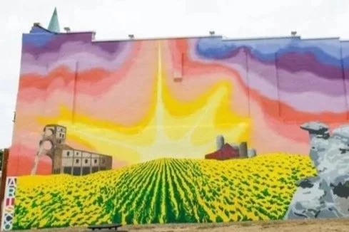 Colorful mural featuring a vibrant sunset sky, a bright yellow field of flowers, and some buildings and rocks.
