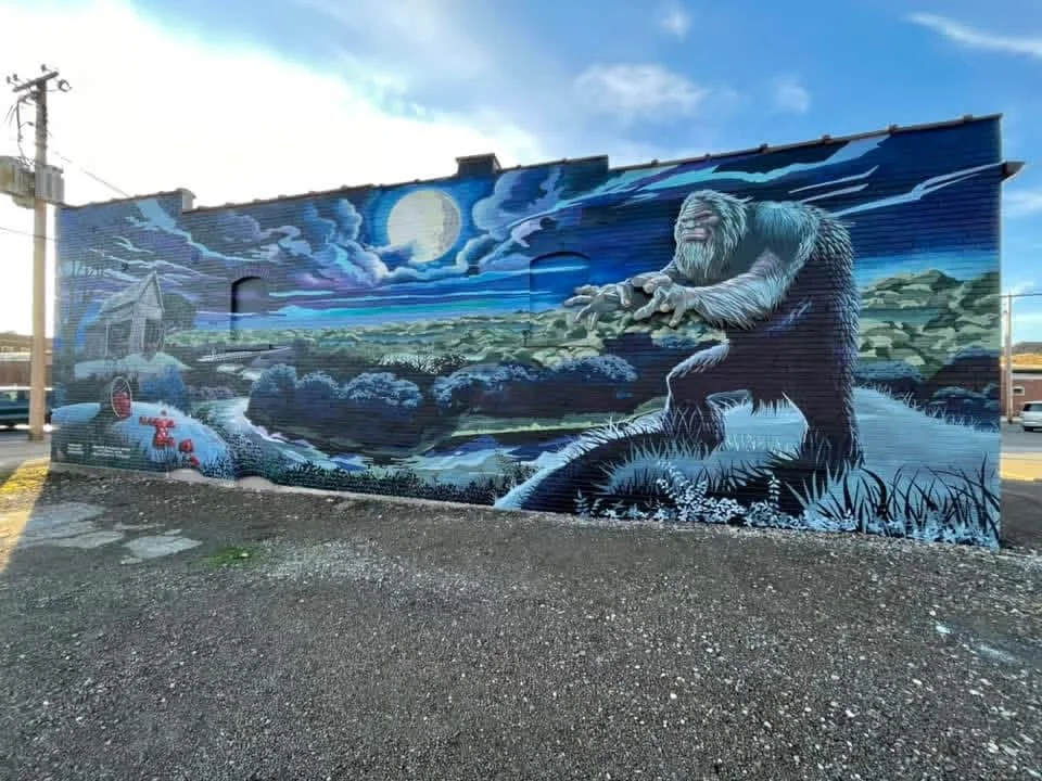 Mural of Bigfoot reaching towards the moonlit sky, with a creek and trees in the background, on a brick building wall.