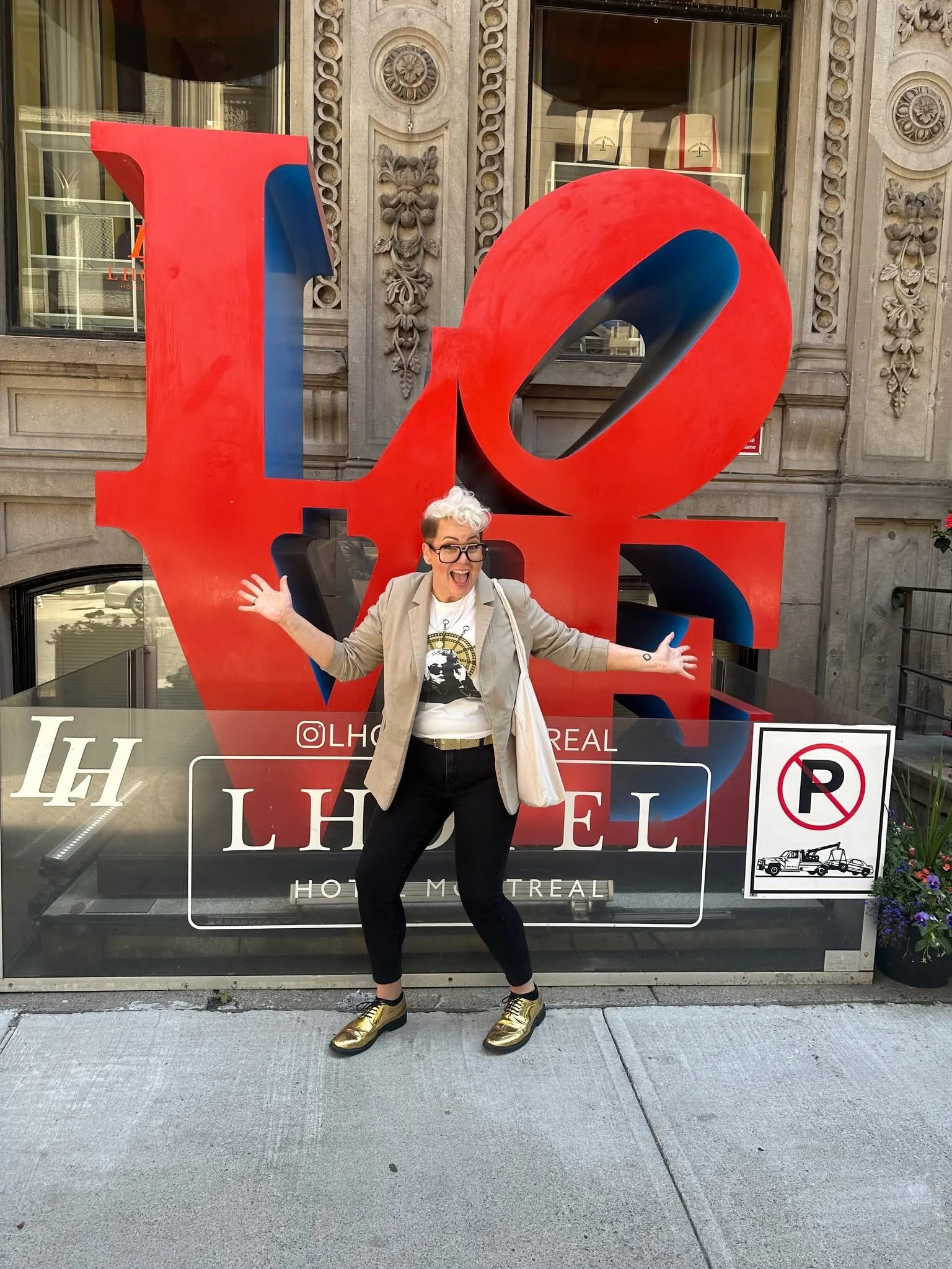 A person with short, curly white hair and glasses stands in front of a large red and blue 'LOVE' sculpture outside a building. They are smiling, wearing a beige blazer over a graphic t-shirt, black pants, and gold shoes, with their arms raised and spread wide.