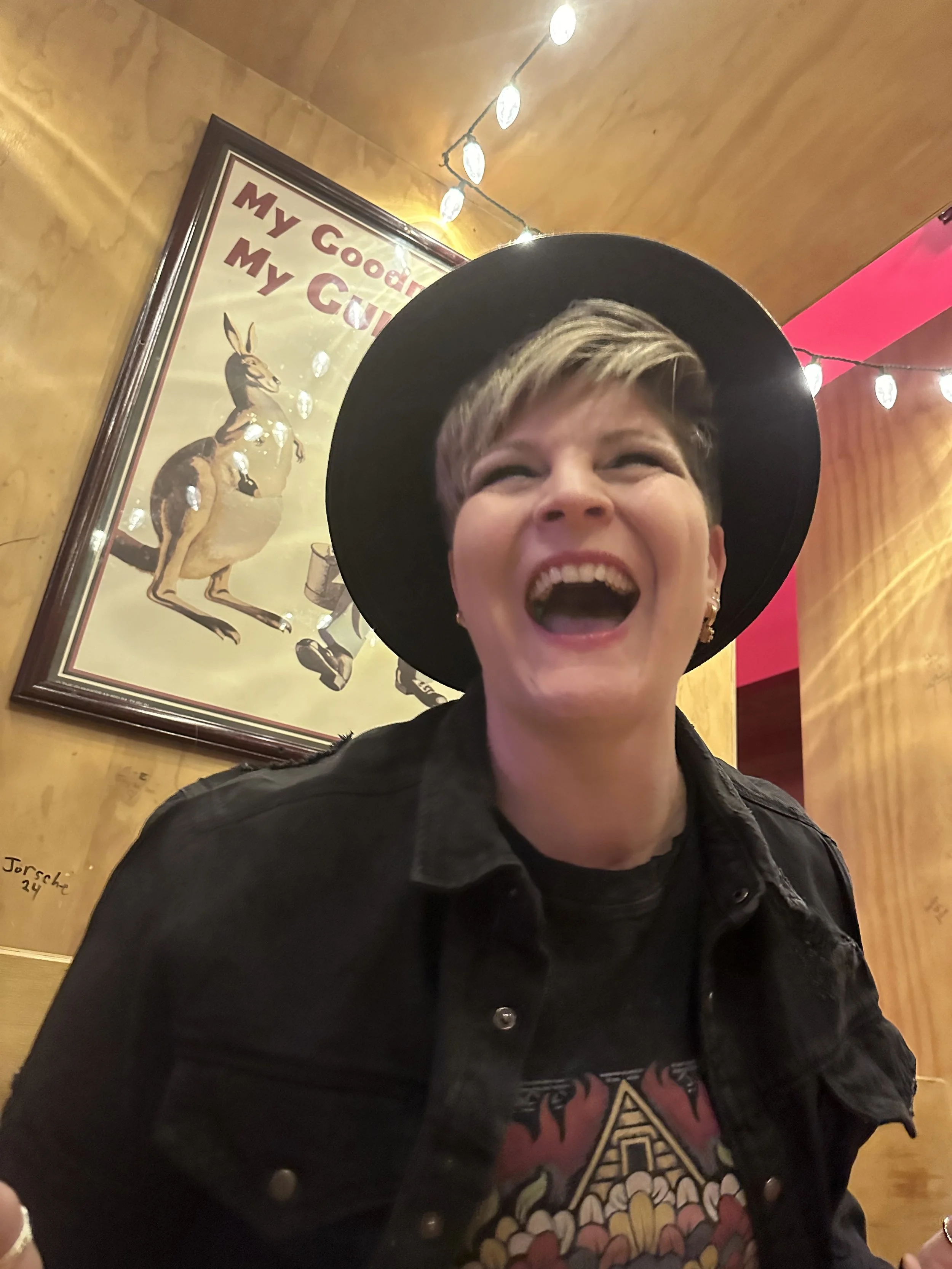 A woman with short blonde hair, wearing a black wide-brimmed hat, black jacket, and a graphic T-shirt, is laughing with her mouth open inside a restaurant or café with warm-colored walls and string lights. There is a framed picture of a kangaroo on the wall behind her.