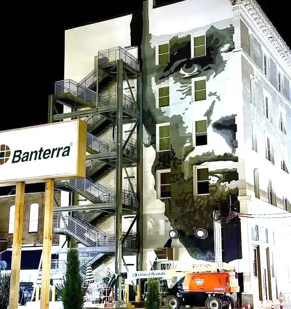 A building with a large mural of a person's face painted on it, featuring multiple windows and an external fire escape. A sign for Banterra and equipment are visible in front.