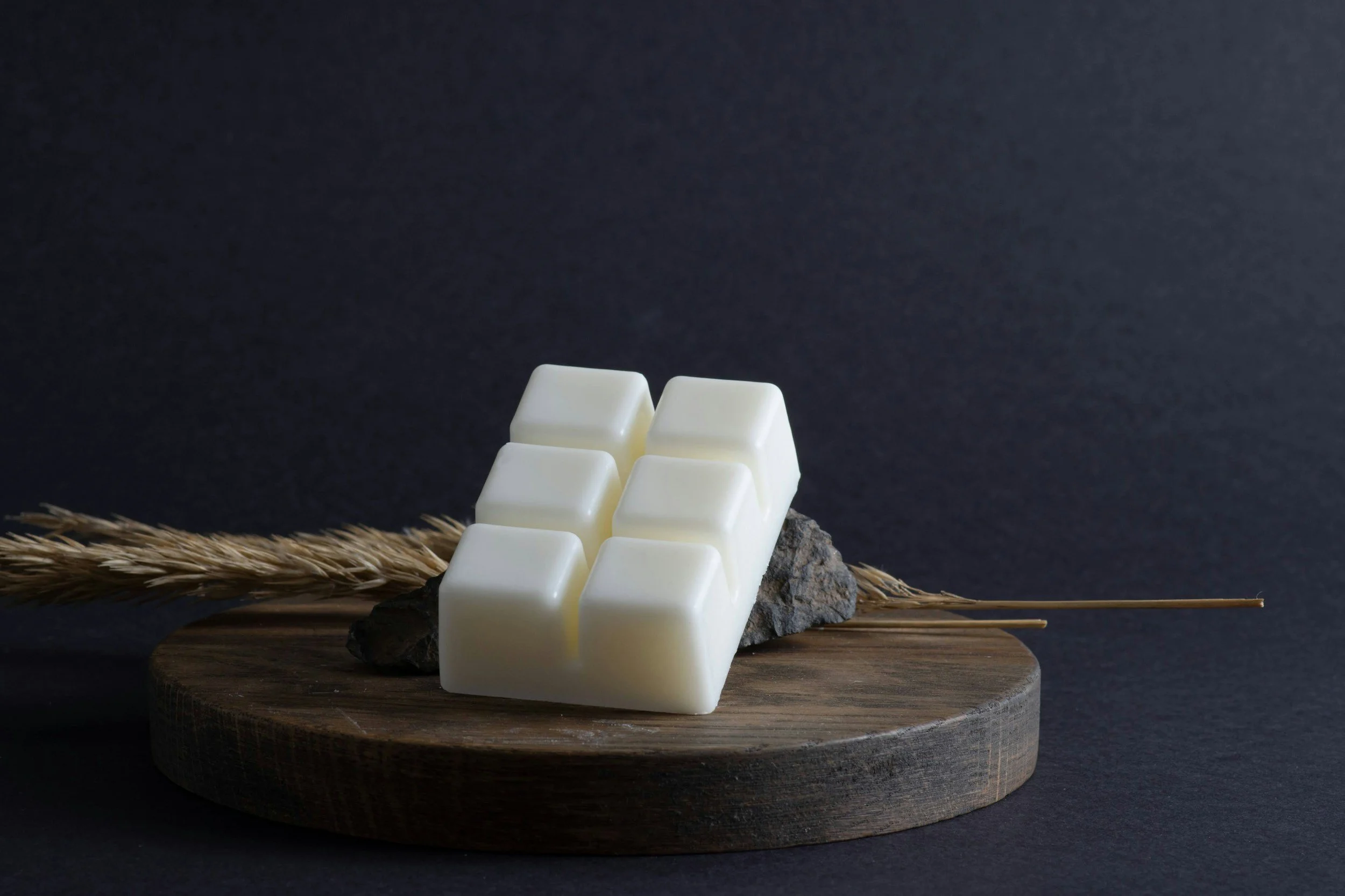 White chocolate bars arranged on a small rock, placed on a wooden platform with dried wheat stalks beside them, against a dark background.
