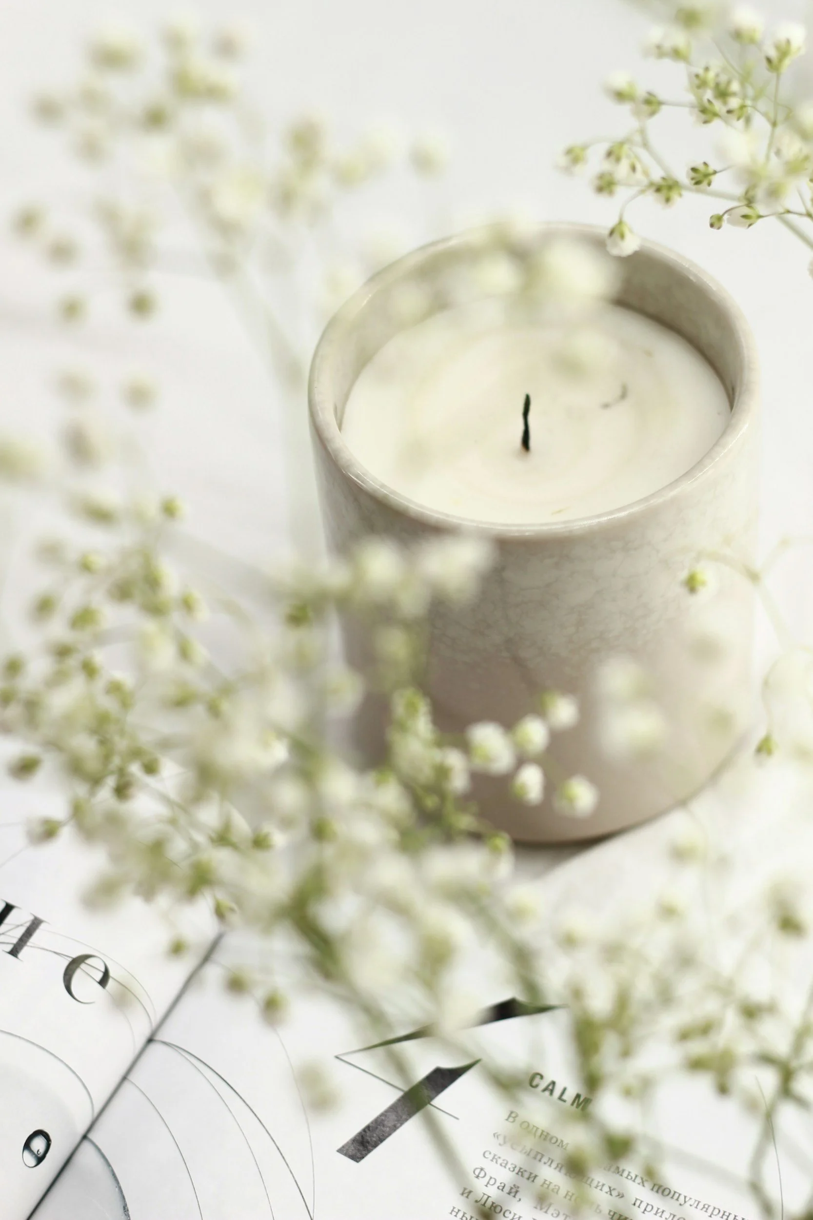 A white candle in a ceramic holder with a black wick, surrounded by delicate white flowers and an open book with printed text.