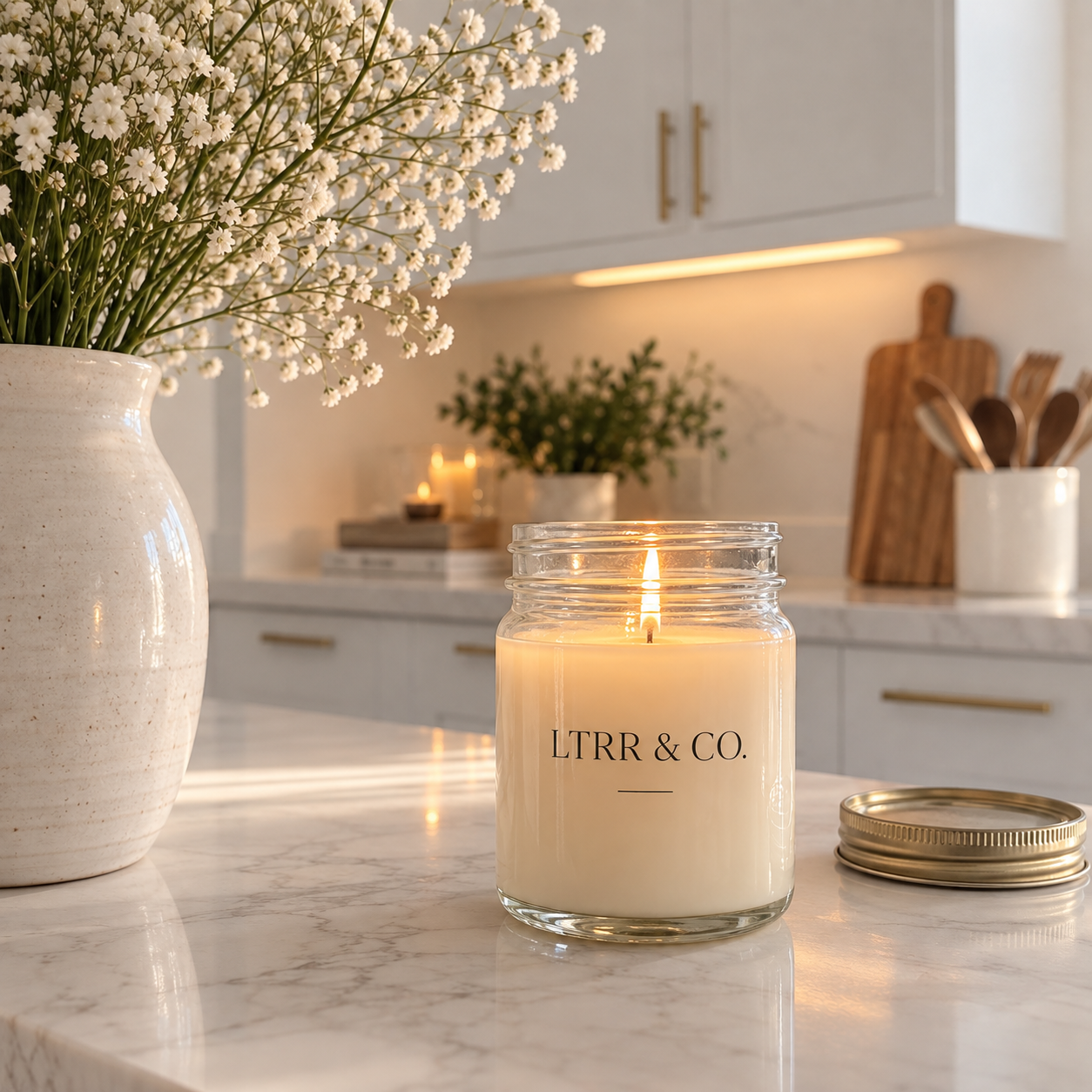 A lit candle in a glass jar with the label 'LTRR & CO.' on a marble kitchen countertop, with a white vase holding white flowers to the left and kitchen utensils and plants in the background.