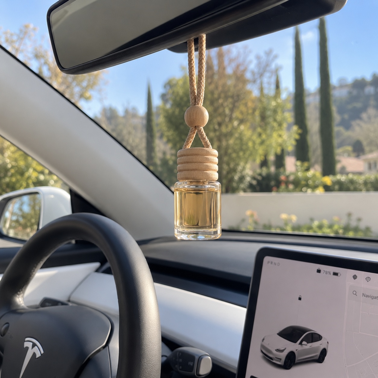 Interior of a Tesla car with a hanging air freshener, a view of the dashboard, and a rearview mirror against a sunny outdoor background with trees and houses.