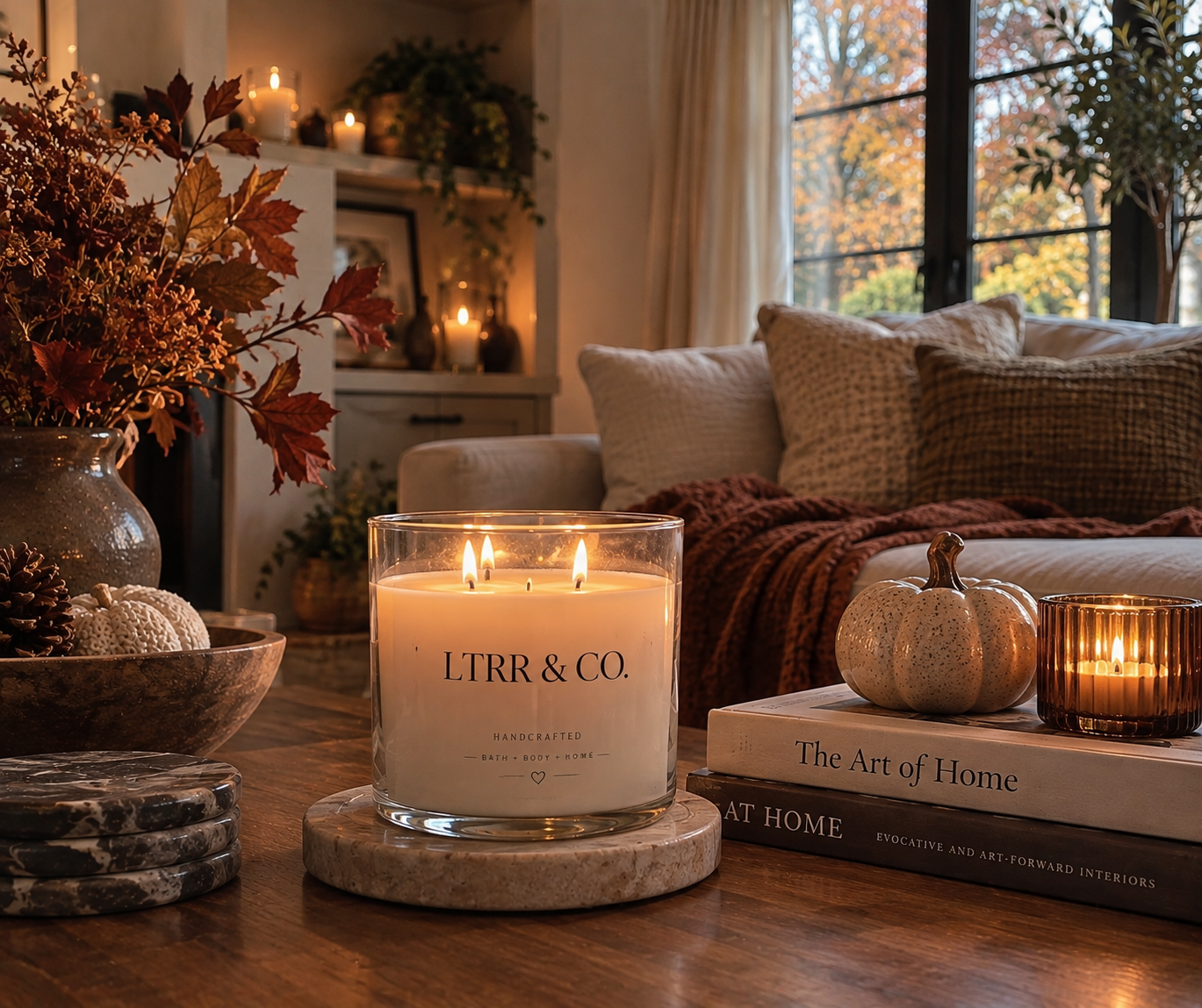 Living room decorated with candles, books, and autumn-themed decor, including pumpkins and dried leaves, with natural light coming through large windows.