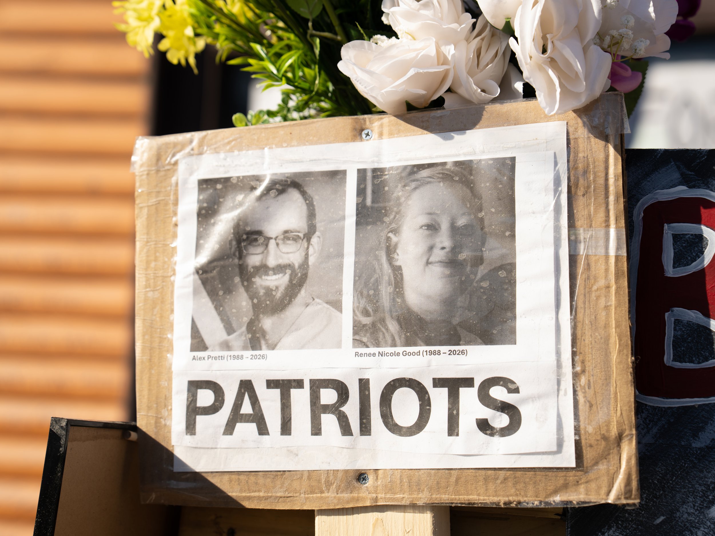 A memorial display with black and white photographs of Alex Pretti and Renee Nicole Good, their birth and death years, and white flowers on top, with a sign that says "Patriots."