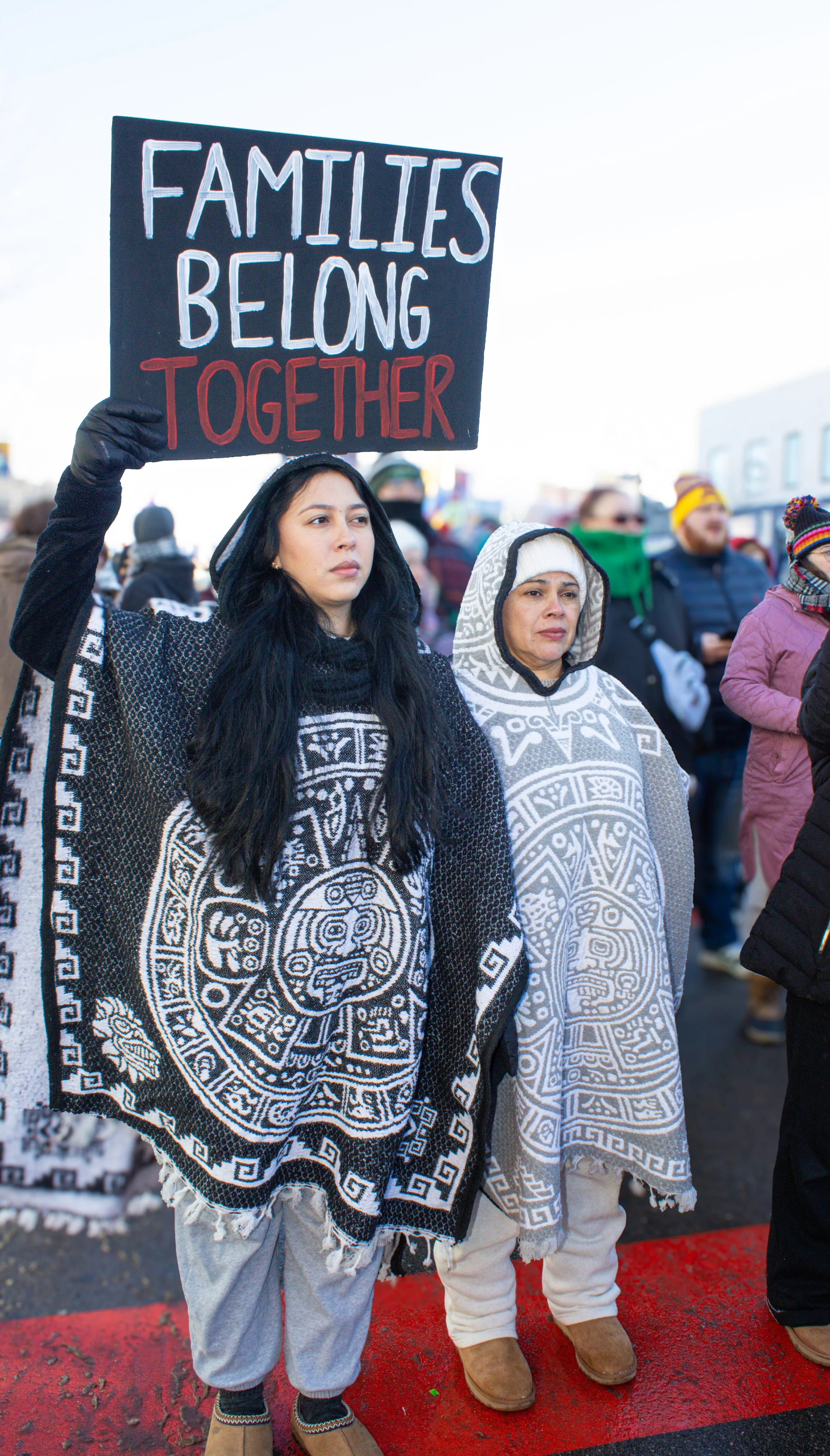 People participating in a protest or march, holding a sign that reads 'FAMILIES BELONG TOGETHER,' outdoors with a crowd.