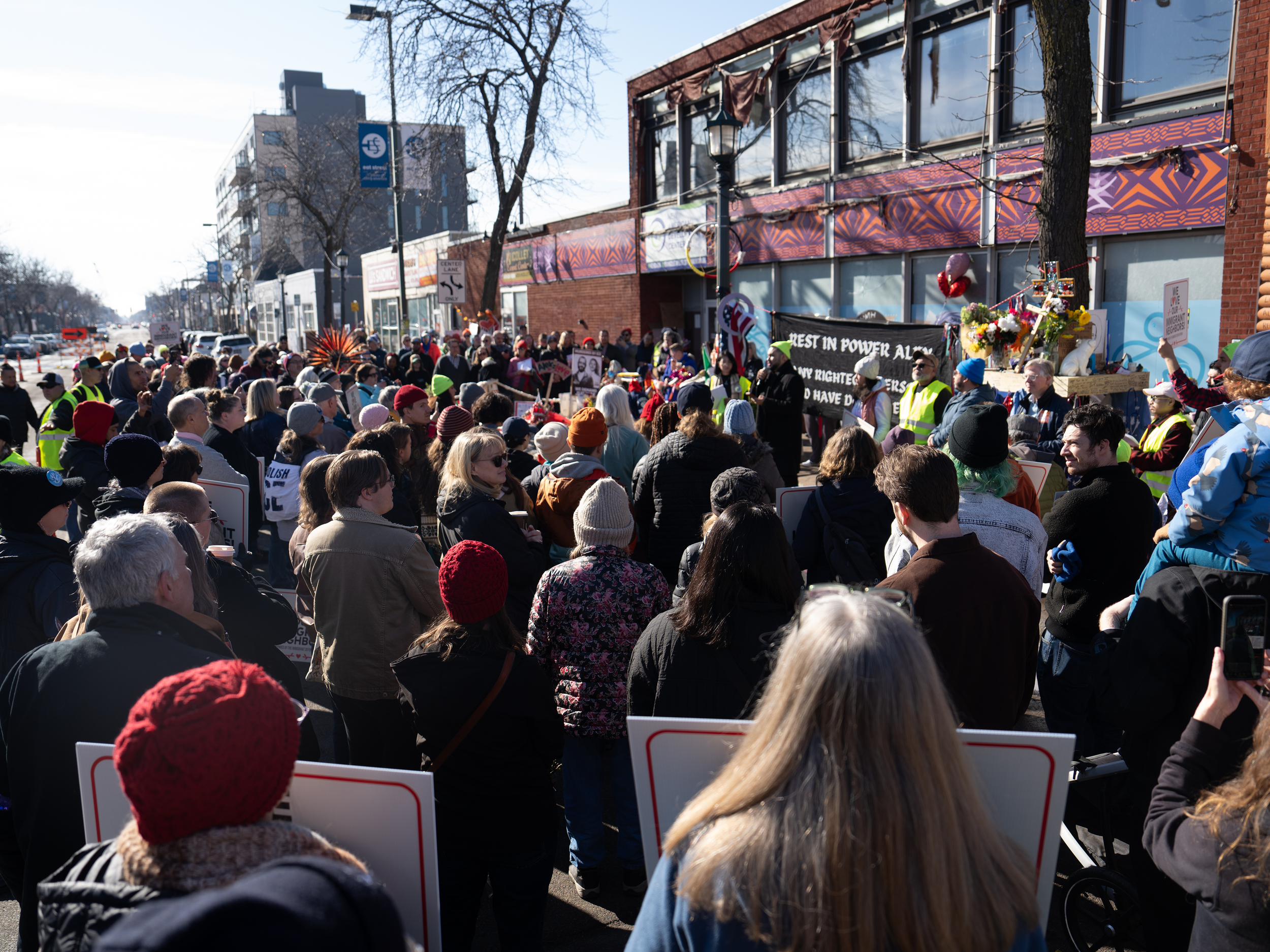 Crowd standing on the street during a protest or rally, with some holding signs and banners, in front of buildings with some trees and a clear blue sky.