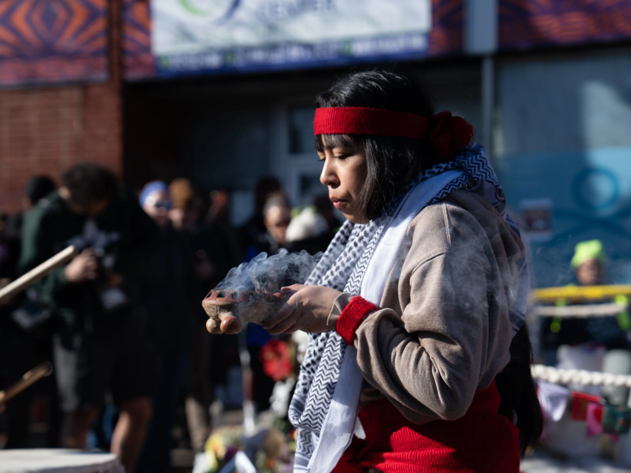 A young woman with dark hair, wearing a red headband and a scarf, holds a rectangular object in her hands while standing among a crowd outdoors on a sunny day. The background includes people and colorful decorations or displays.