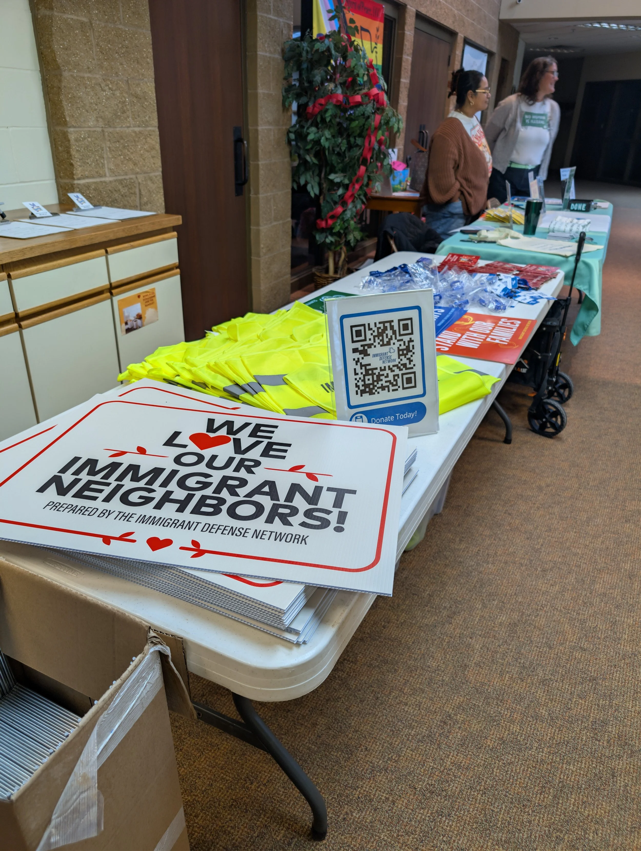 Table with flyers and signs promoting immigrant neighborhood awareness and donations, volunteers behind the table, holiday decorations, and QR code for donations.