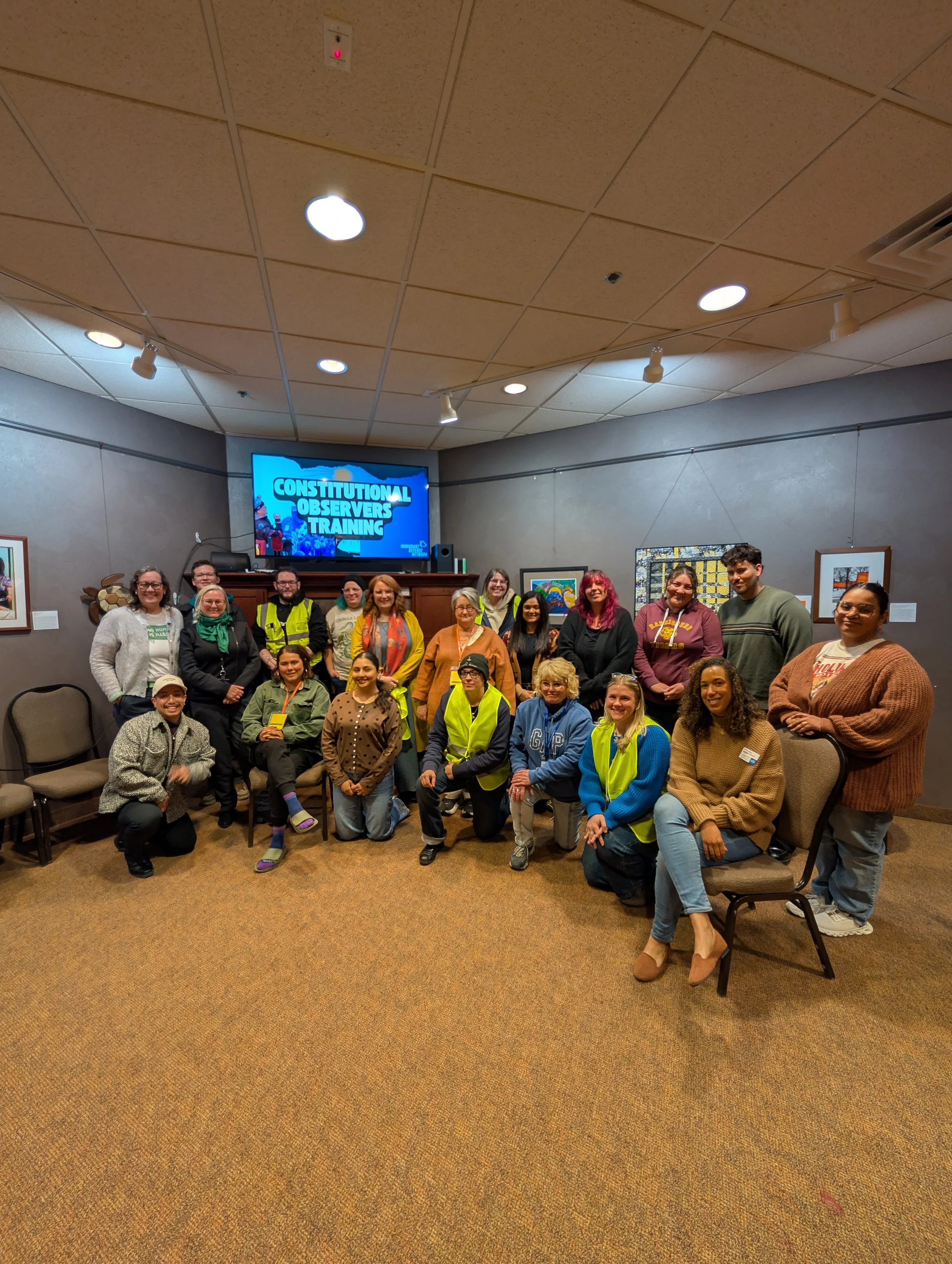 Group of diverse people standing and sitting in a room with artwork on the walls, in front of a large television screen displaying 'Constitutional Observers Training'.