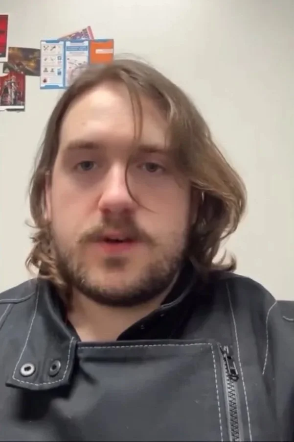 A man with shoulder-length brown hair and a beard wearing a black leather jacket, sitting indoors with a plain wall and some posters in the background.