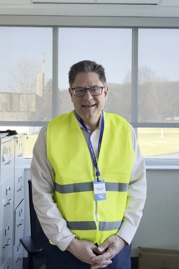 A man wearing glasses and a white shirt with a yellow safety vest standing indoors near large windows with an outdoor view.