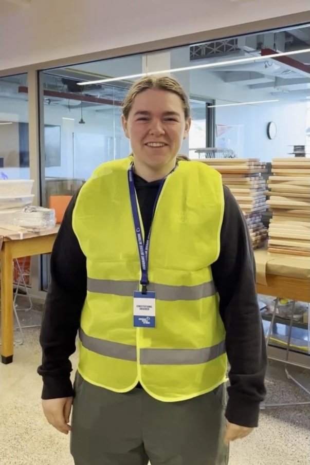 A young woman wearing a yellow safety vest and an ID badge standing inside a room with stacks of wood or paper.