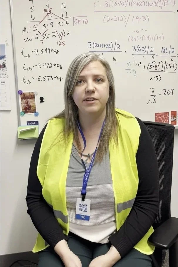 A woman wearing a yellow safety vest sitting in a black mesh office chair in front of a whiteboard filled with mathematical equations and notes, with a lanyard around her neck.