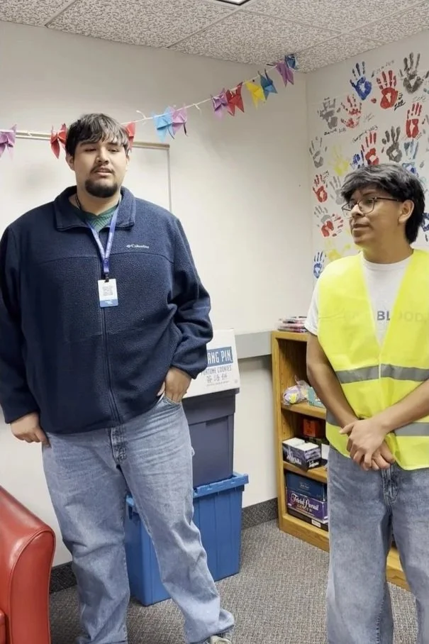 Two young men standing indoors in a room decorated with colorful paper banners and handprints on the wall. One is wearing a dark jacket with a lanyard, and the other is wearing a yellow safety vest over a white T-shirt.