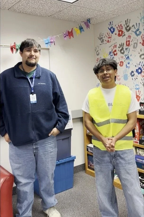 Two young men standing in a room decorated with colorful paper flags and handprints on the wall. One is wearing a dark jacket and jeans, and the other is wearing glasses, a white t-shirt, and a yellow safety vest.