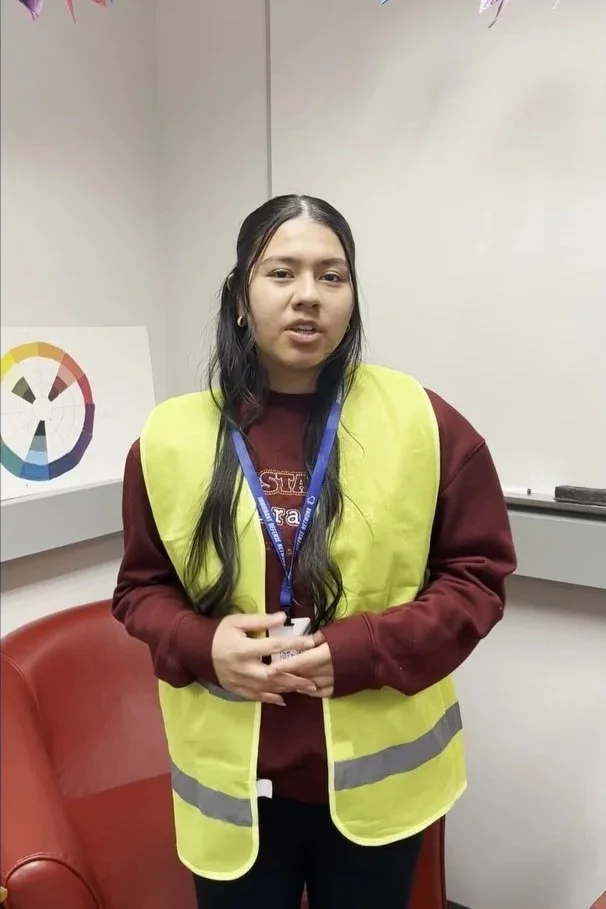 A woman wearing a yellow safety vest standing in an office room with a whiteboard and a colorful chart on the wall.