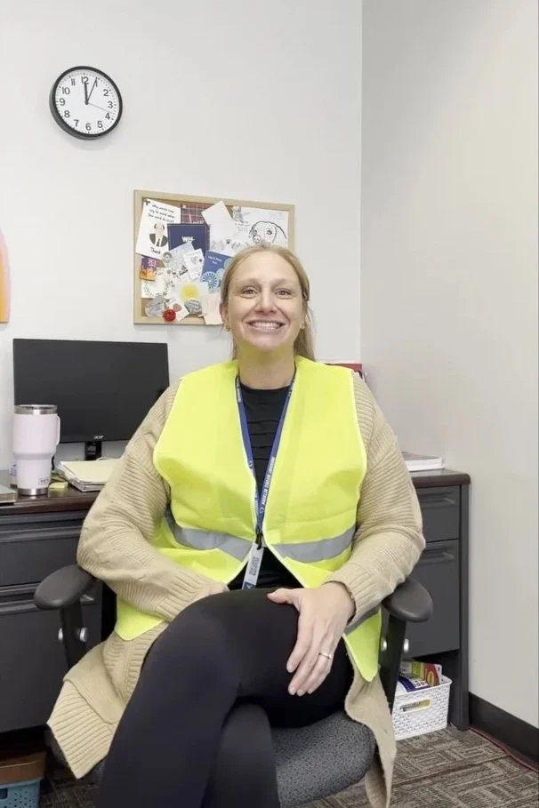 A woman sitting in an office chair, smiling, wearing a yellow safety vest and casual beige cardigan, with a computer and office supplies behind her in a workplace.