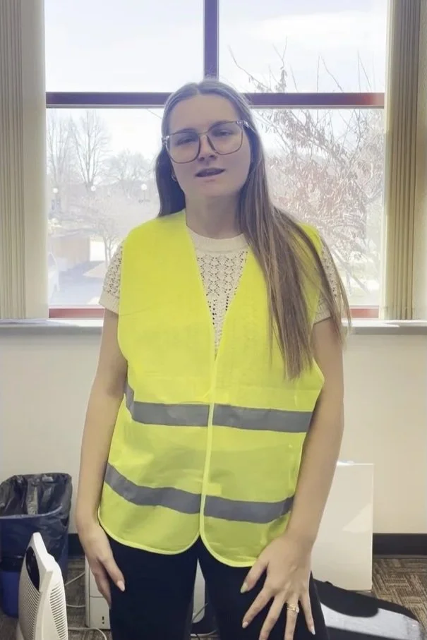 Young woman wearing glasses and a bright yellow safety vest, standing indoors near a window.