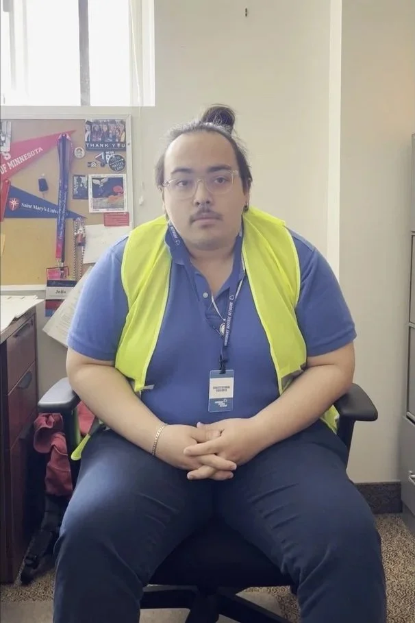 A man with glasses and a ponytail wearing a blue shirt and a yellow safety vest, sitting in an office chair with hands clasped on his lap.