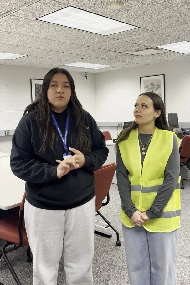 Two women standing in an office or classroom setting, one wearing a black sweatshirt and light-colored sweatpants, the other wearing a yellow safety vest and gray pants, with computers and framed pictures on the wall in the background.