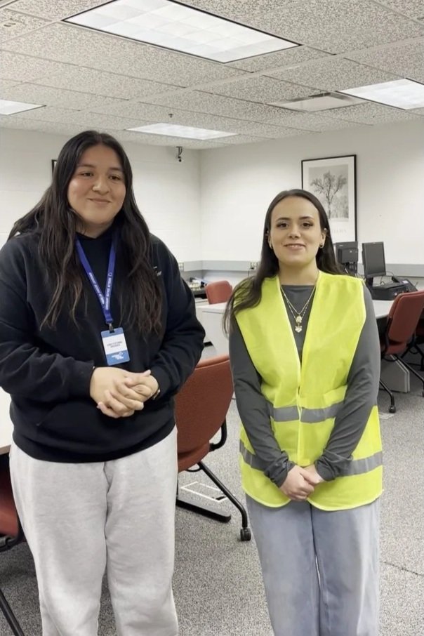 Two women standing in an office with computers and chairs behind them. One woman is wearing a black hoodie and light-colored sweatpants; the other is wearing a gray long sleeve shirt with a bright yellow safety vest and light-colored jeans.