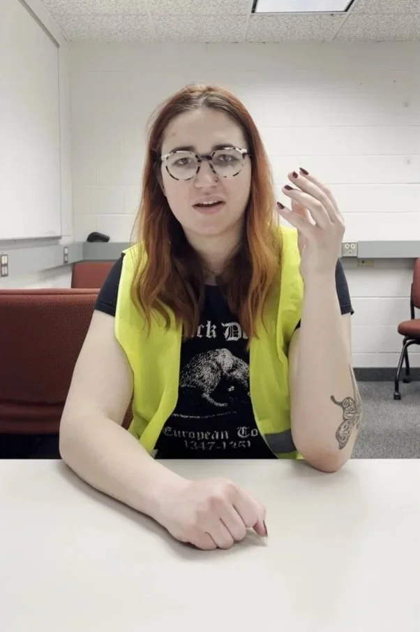 A woman with long red hair and glasses sitting at a table in a classroom or conference room, wearing a black T-shirt, a yellow vest, and showing a tattoo on her left arm.