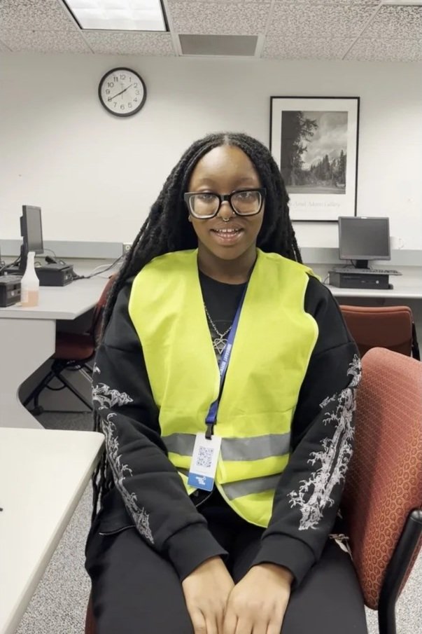Young woman with glasses and dreadlocks sitting in front of computers, wearing a yellow safety vest and a black hoodie with white embroidery, in an office or classroom setting.