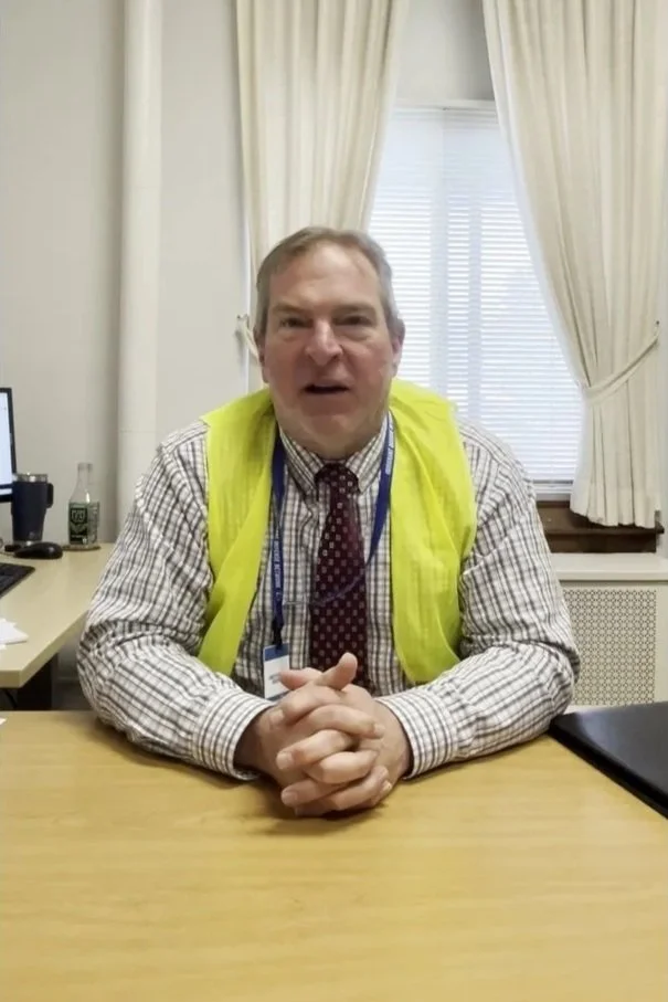 Middle-aged man sitting at a desk, wearing a yellow safety vest, checkered shirt, and red tie with white dots, in a room with beige curtains and a window with blinds.