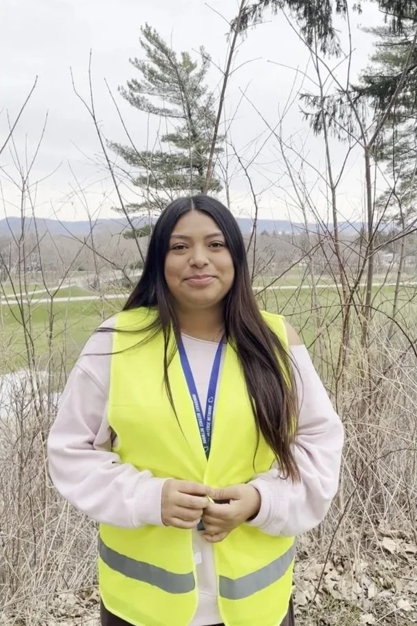 A woman wearing a yellow high-visibility safety vest standing outdoors in front of leafless trees and a landscape of grass and distant mountains, overcast sky.