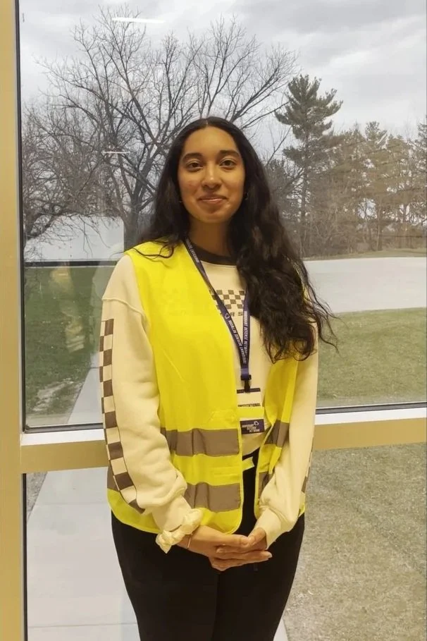 A young woman standing indoors in front of a large window with a view of a tree and grass outside. She is wearing a yellow safety vest over a beige sweatshirt with patterned stripes on the sleeves.