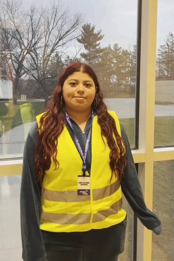 A woman with long curly red hair wearing a yellow safety vest and an ID badge standing indoors near a window, with trees and a cloudy sky outside.