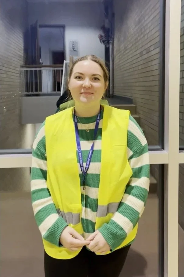 Young woman wearing a yellow safety vest and a lanyard standing indoors near large windows.