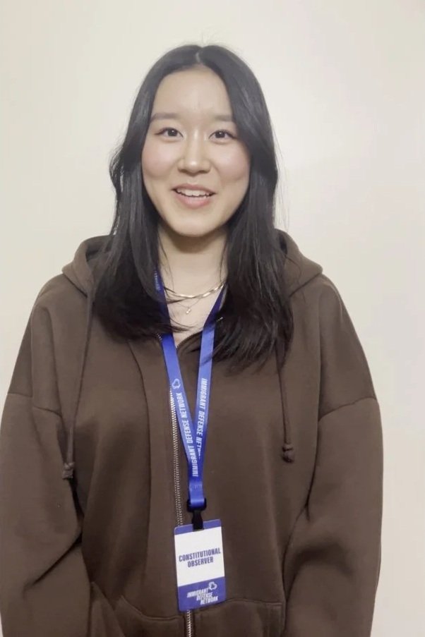 A young woman with black hair wearing a brown hoodie and a blue lanyard with a badge that says 'Constitutional Observer' standing against a plain off-white wall.