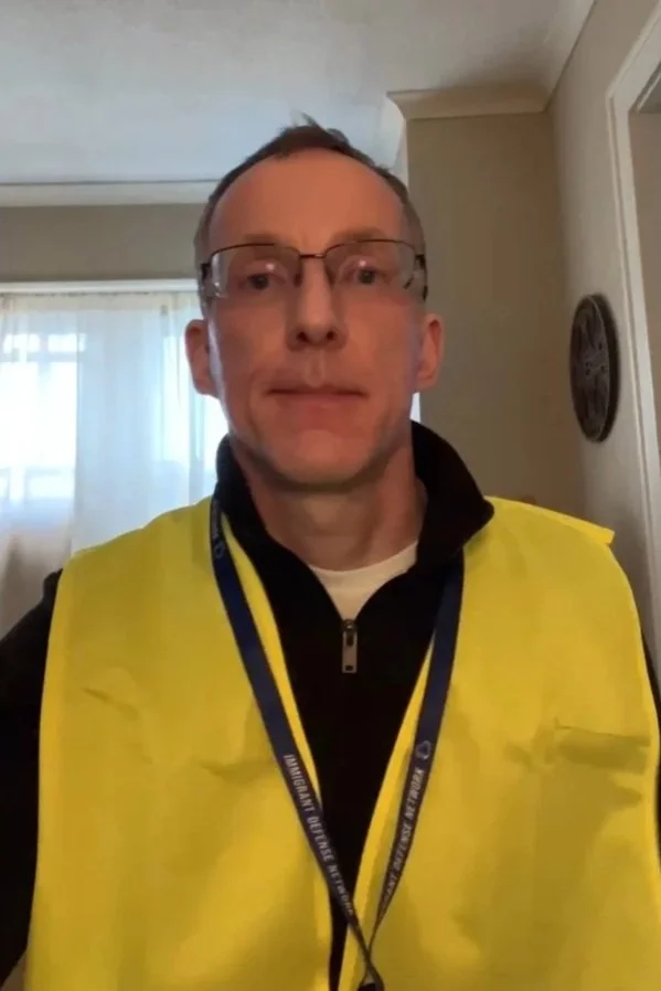 A middle-aged man wearing glasses, a yellow safety vest over a black zip-up jacket, and a lanyard around his neck, standing indoors in a room with light-colored walls and a window with sheer curtains.