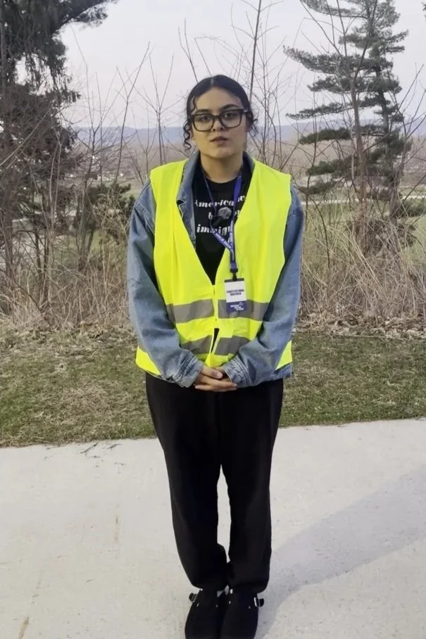 A young woman with glasses wearing a yellow safety vest, black pants, and black shoes standing outdoors on a sidewalk with trees and dry bushes in the background.