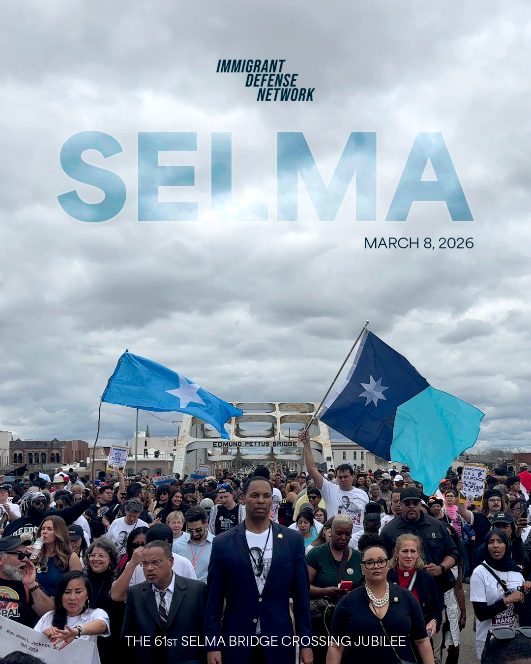Crowd of people gathered for the 61st Selma Bridge Crossing Jubilee, with two flags being held up, under a cloudy sky, in Selma, Alabama.