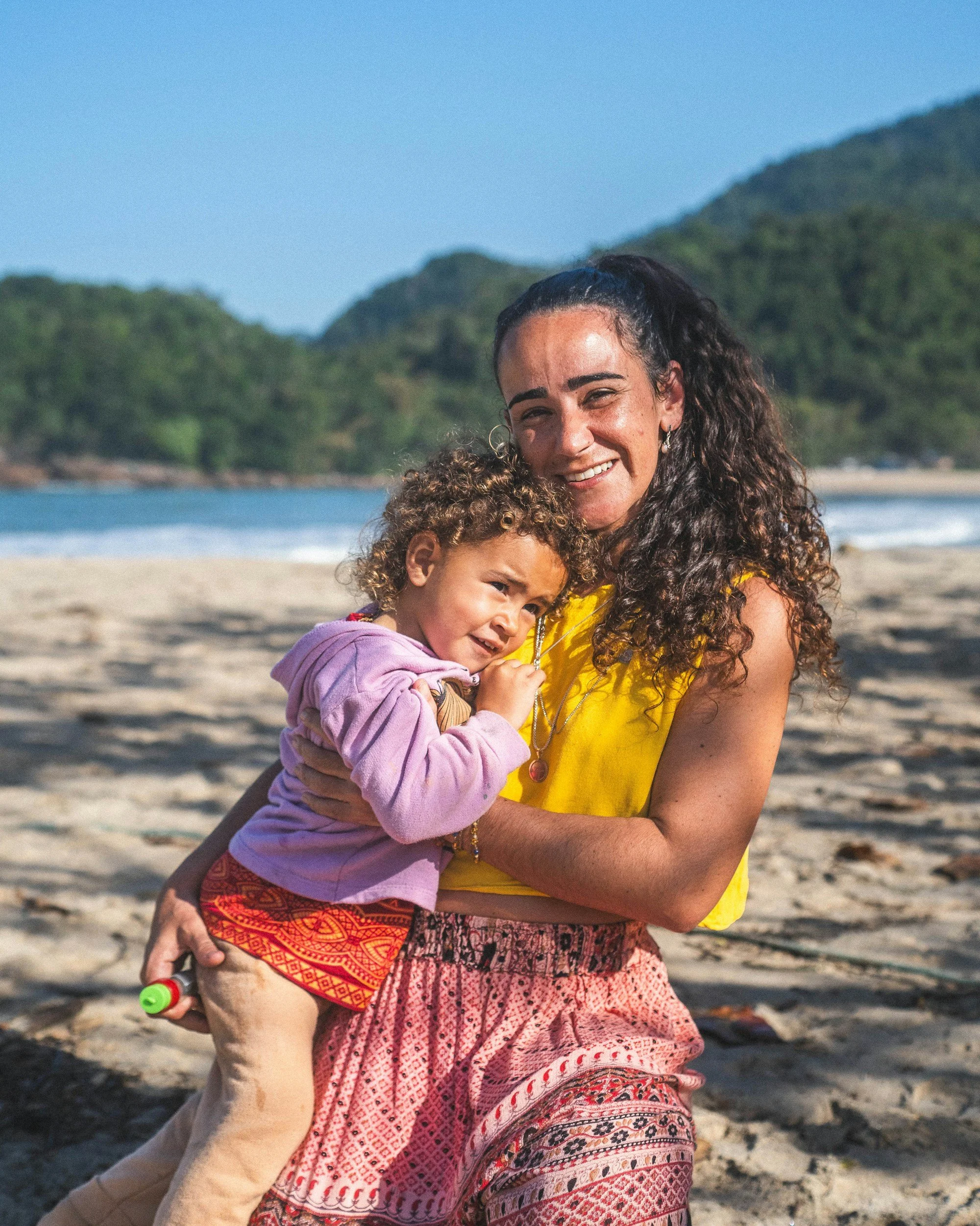 A woman with curly hair wearing a yellow top and patterned skirt holding a young girl with curly hair wearing a pink jacket at the beach with ocean and hills in the background.