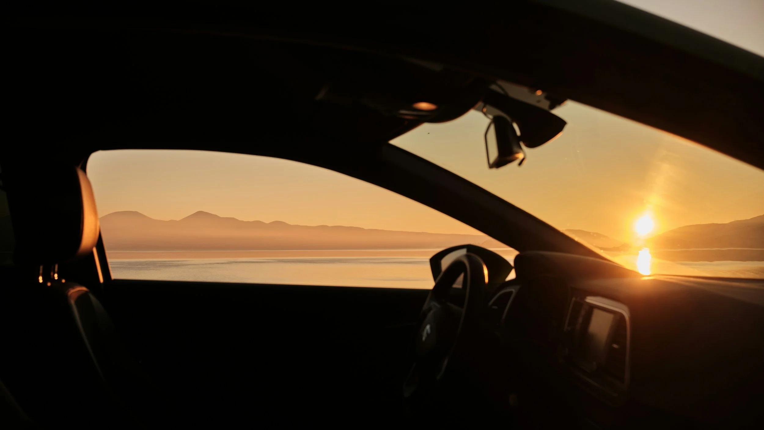 Intérieur d'une voiture, vue sur un coucher de soleil sur un lac avec des montagnes en arrière-plan.