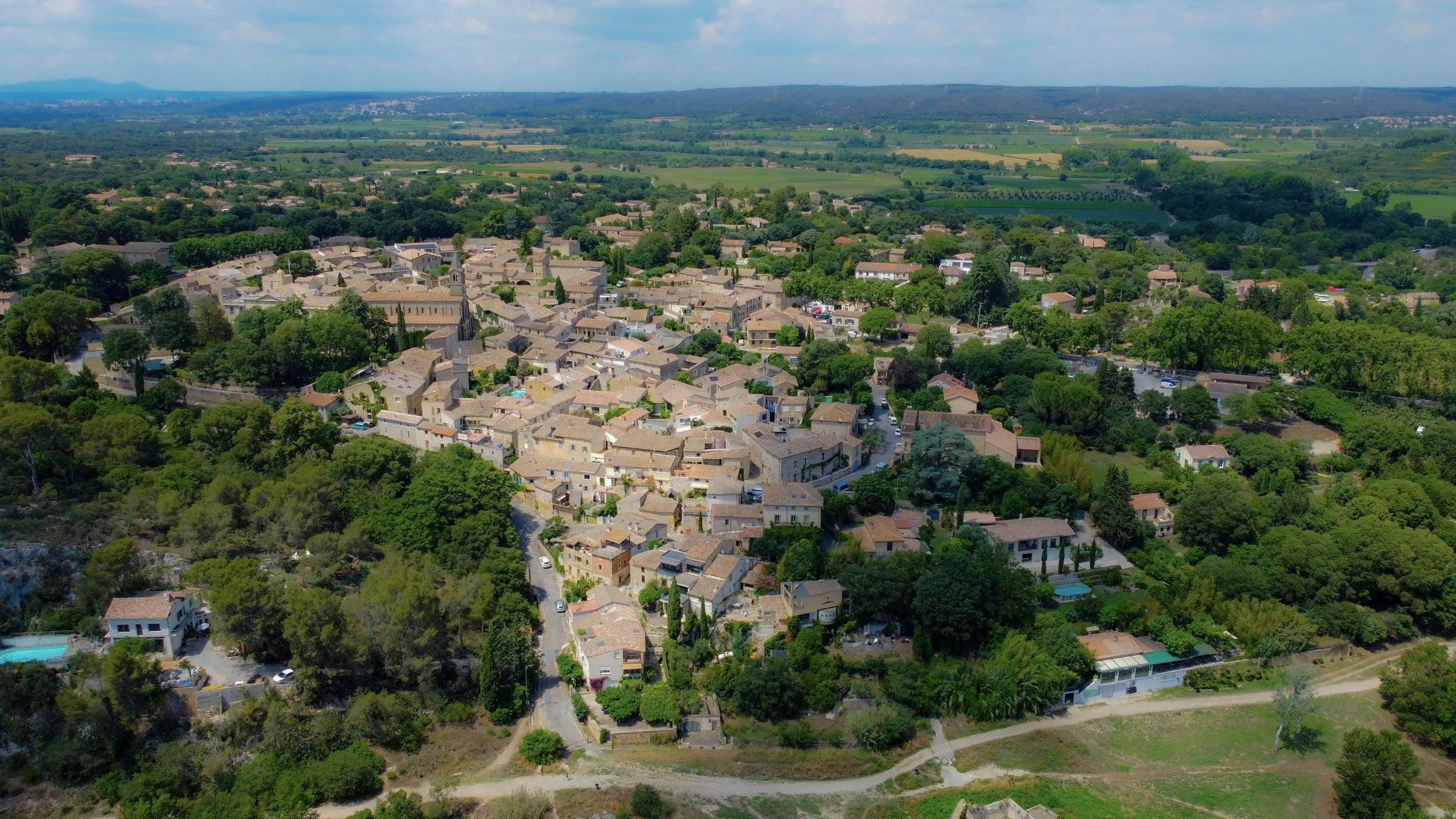 Vue aérienne d'un village entouré de verdure avec plusieurs maisons, églises, routes et jardins, en arrière-plan des champs agricoles et une chaîne de montagnes sous un ciel partiellement nuageux.