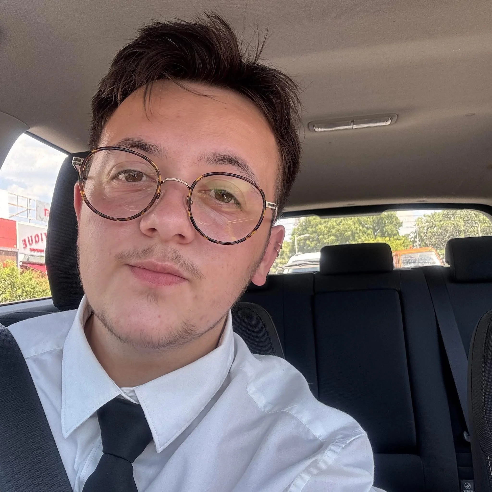 Le chauffeur portant des lunettes rondes, chemise blanche et veste sombre, assis dans une voiture avec un ciel clair visible par la fenêtre.