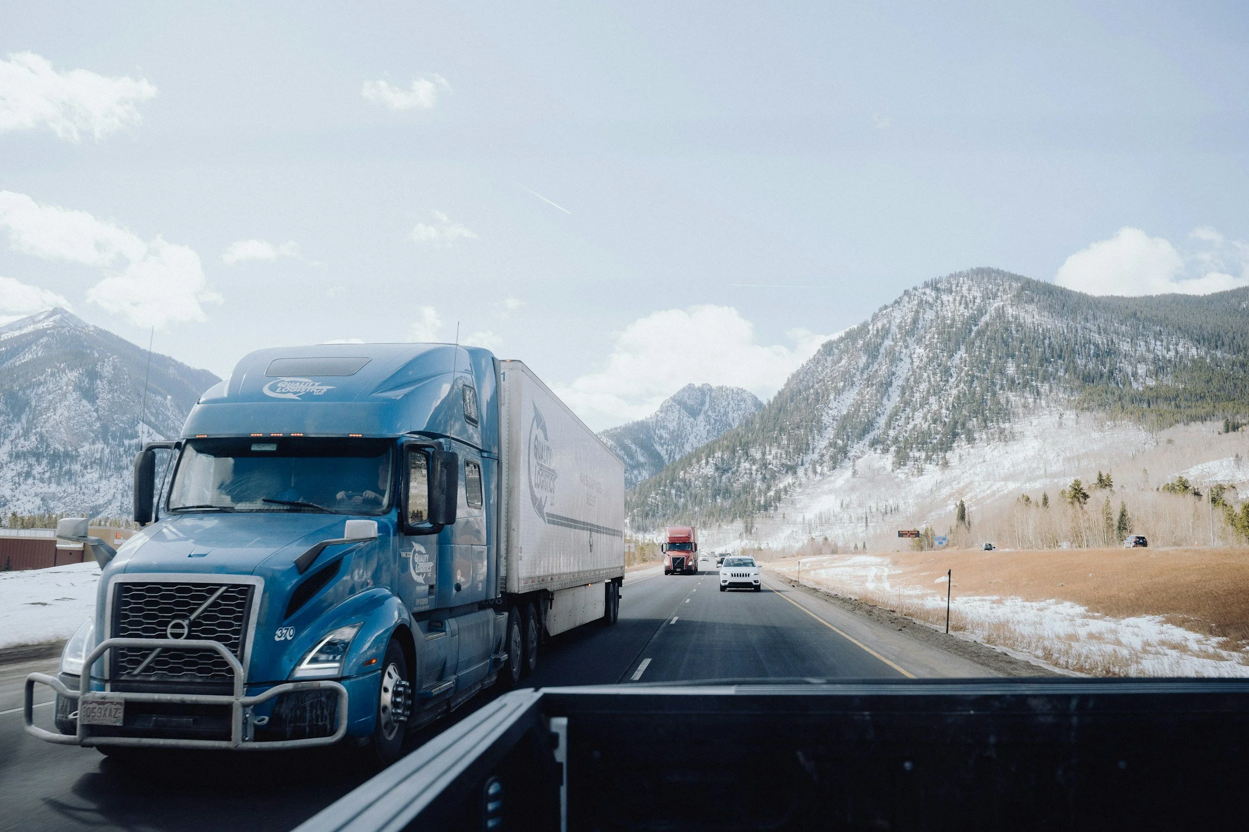 A blue semi-truck driving on a highway with mountains and partly cloudy sky in the background.