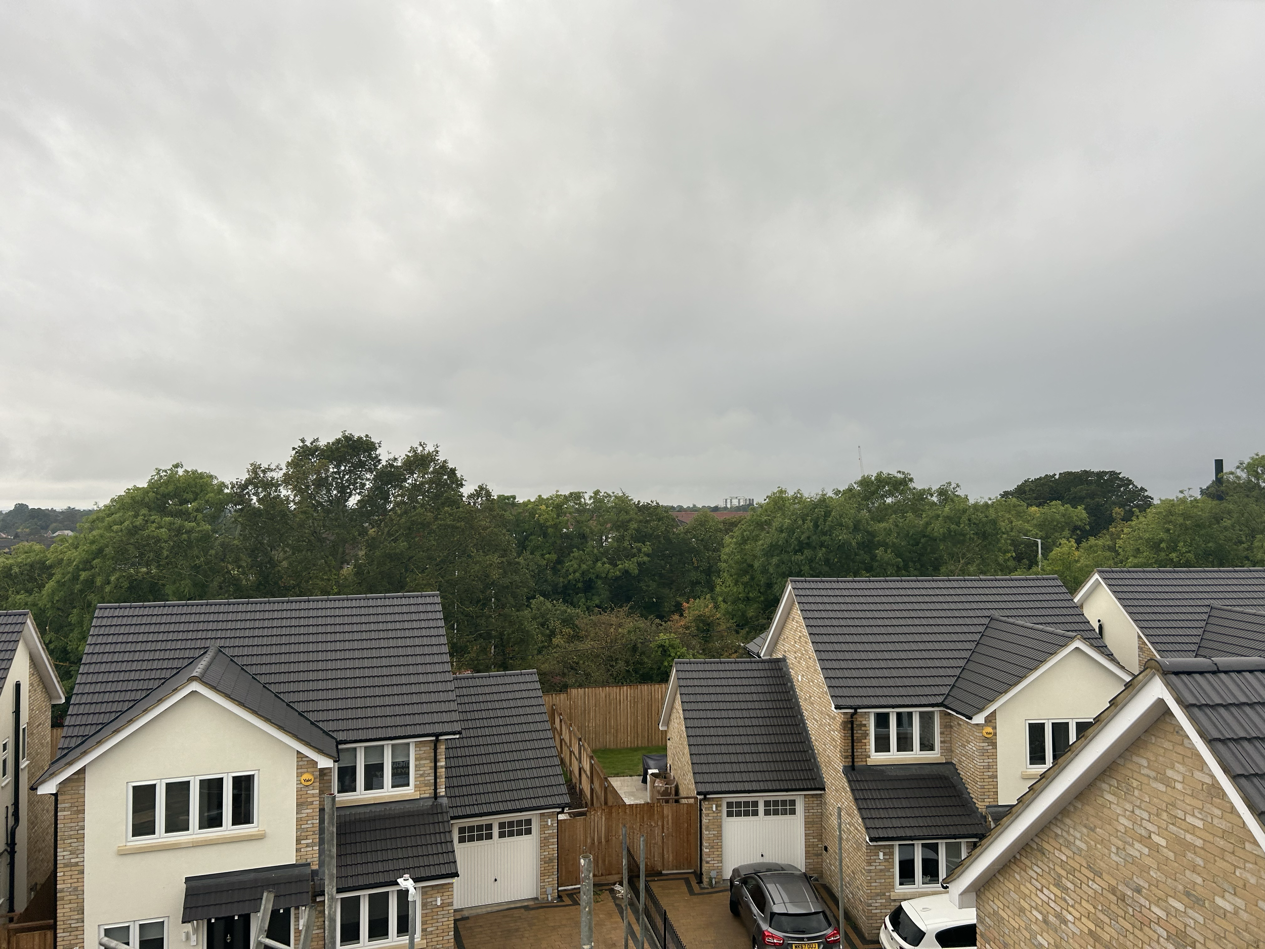 Residential neighborhood with several houses featuring brick and siding exteriors, dark gray tiled roofs, and small yards. There are cars parked in driveways and the tops of trees and a cloudy sky in the background.