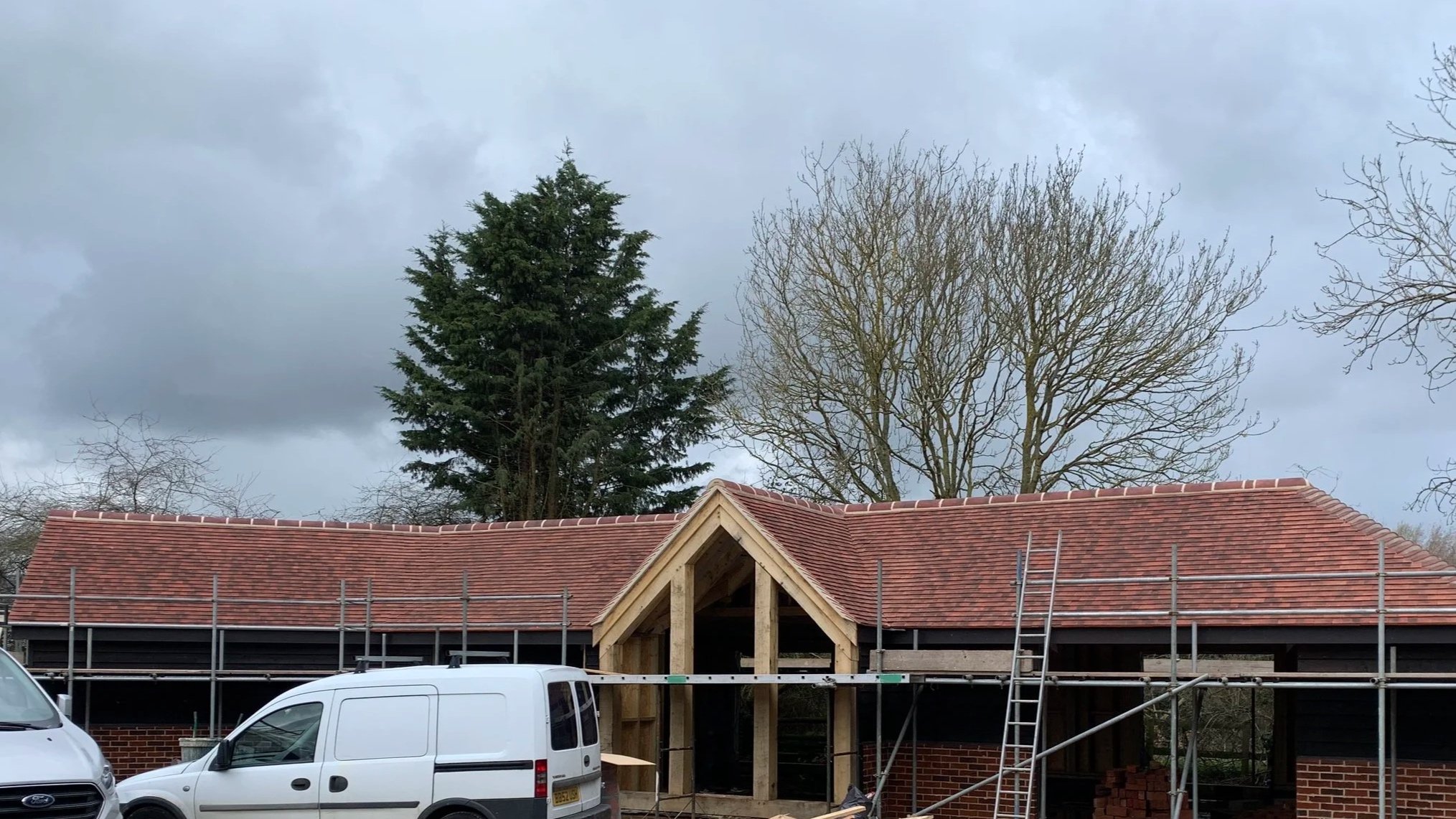 A house under construction with a red tiled roof and scaffolding around it. Trees with bare branches and a cloudy sky in the background.