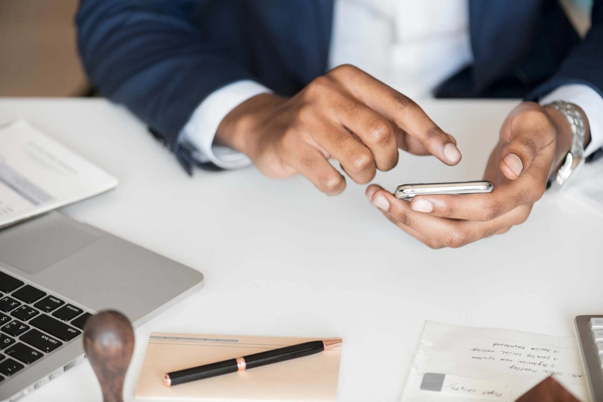 Person in a business suit using a smartphone at a desk with a laptop, pen, and notepad.