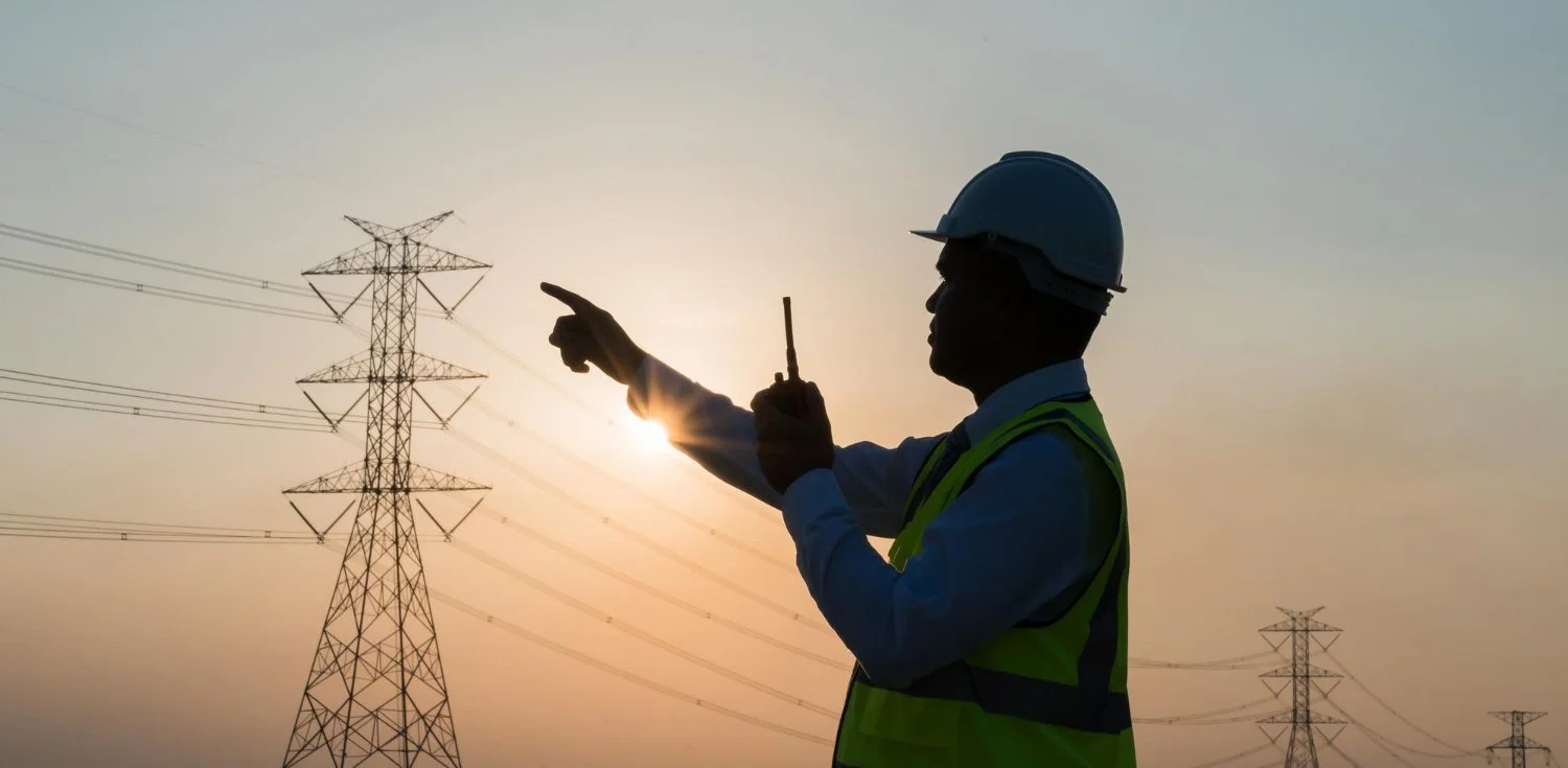 A silhouette of a utility worker wearing a hard hat and safety vest, pointing at high-voltage power lines against the sunset.