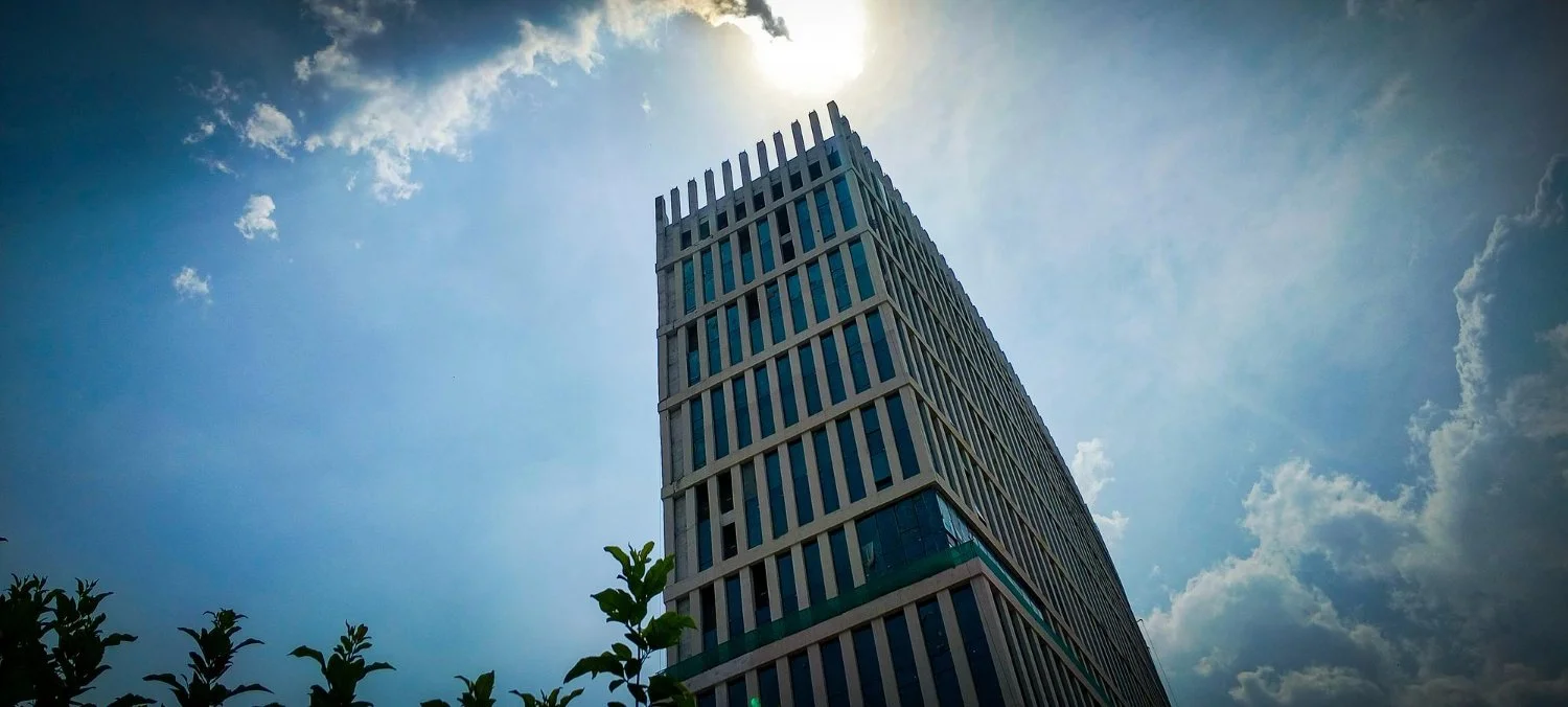 A tall modern skyscraper with a pattern of vertical and diagonal windows, taken from a low angle with a partly cloudy sky and bright sun in the background.